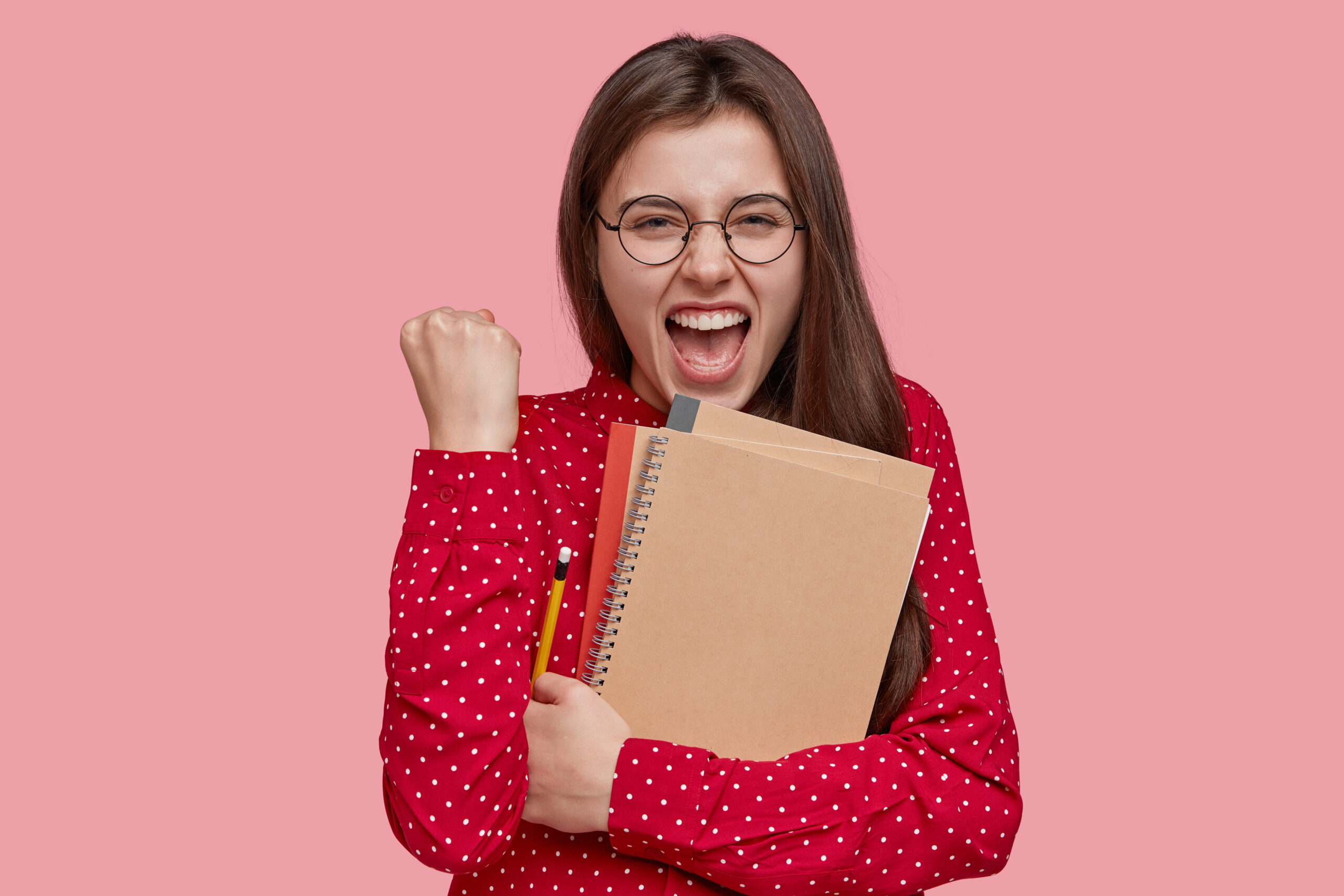 positive happy teacher clenches fist with joyful expression, holds spiral notepad, writes notes in notepad, dressed in red shirt, exclaims with happiness, models over pink background. studying concept